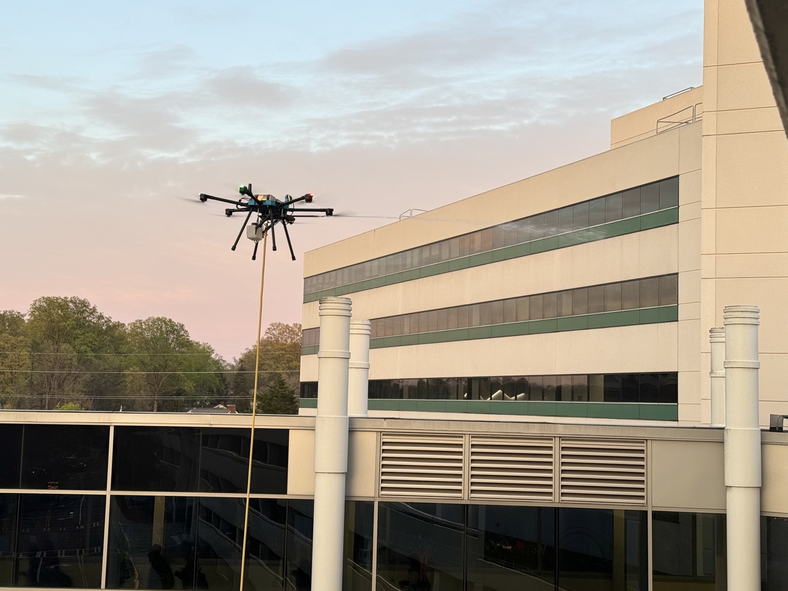 Cleaning drone hovering beside a commercial building at dusk with the supply hose running down
